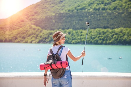 girl traveler makes selfi with an action camera on a mountain lake. She makes a photo for travel blog View from back of the tourist travelerの写真素材
