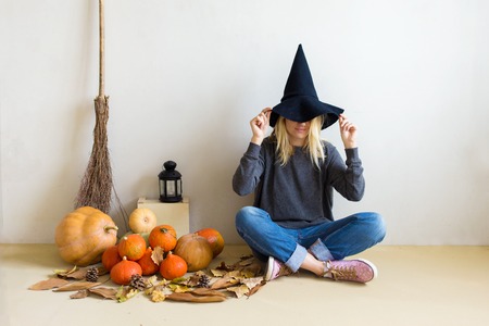 Halloween concept. Beautiful blond girl in a witch hat with pumpkins and broom sitting on the floor against a white wall background Copy spaceの写真素材