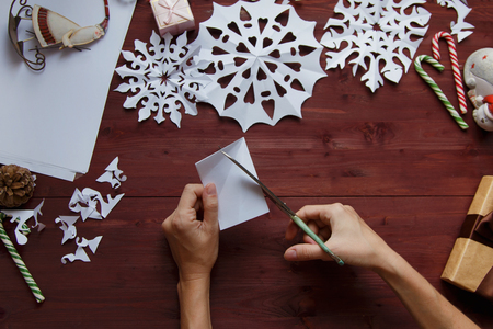 New Year's, Christmas concept. Female hands cut snowflakes from paper. In the background lie gifts, Christmas decorations on a wooden background Flat layの写真素材