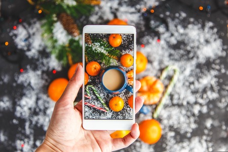 Christmas and New Year's concept. Female hands photographed on a smartphone a New Year's composition of mandarin, spruce branches, snow and a cup of coffee Top viewの写真素材