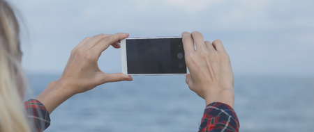beautiful girl takes photo of the sea. Women's hands holding smartphone. The sea is blurred Bannerの写真素材
