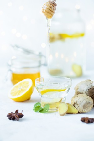 Honey is poured into a ginger drink in a cup. Ginger root, honey in a jar, lemon on a white table.の写真素材
