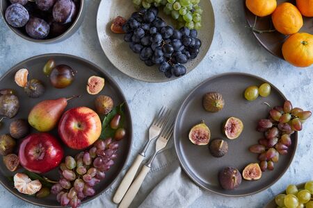 Assortment healthy organic fruits in plates apple grapes fig plums tangerines on light tableの写真素材