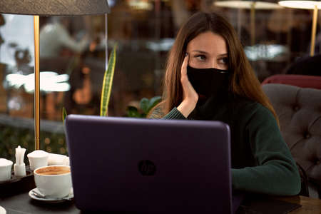 Young brunette woman in medical mask in cafe with laptopの写真素材