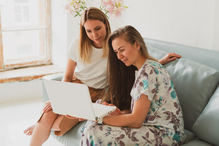 two charming young women chatting at home on the couch, drinking coffee and working on a laptop