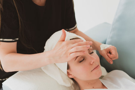 beautician makes facial and massage and cosmetic cream to adorable young woman with a towel on her headの写真素材