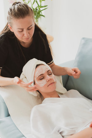 beautician makes facial and massage and cosmetic cream to adorable young woman with a towel on her headの写真素材