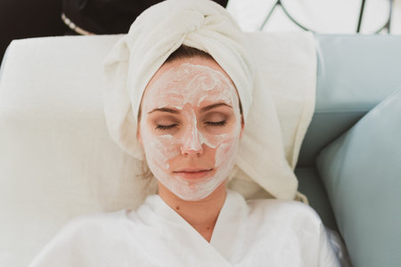 beautician makes facial and massage and cosmetic cream to adorable young woman with a towel on her headの写真素材