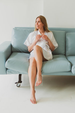 portrait of a young adorable woman in a silk robe with cup of coffee at home on the sofaの写真素材