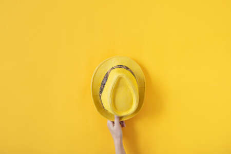 Female hand holding yellow straw hat on yellow background Concept photoの写真素材