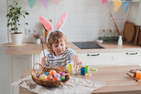 Cute two-year-old boy in bunny ears paint Easter eggs with multi-color paints sitting at the table...