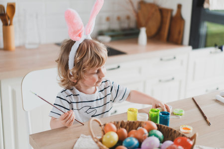 Cute two-year-old boy in bunny ears paint Easter eggs with paints sitting at the table on the...