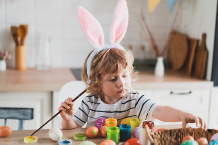 Cute two-year-old boy in bunny ears paint Easter eggs with paints sitting at the table on the kitchenの写真素材