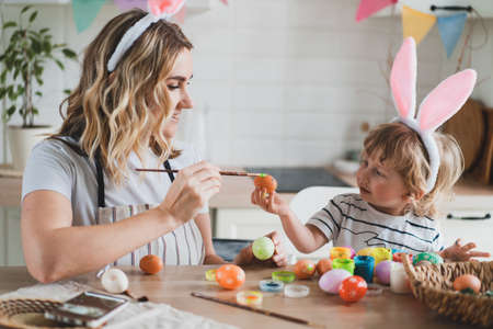 charming woman and her two-year-old son in bunny ears paint Easter eggs with paints sitting at the table on the kitchenの写真素材