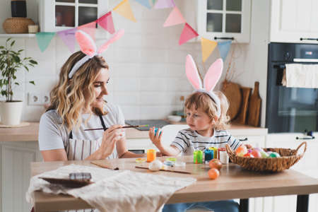 charming woman and her two-year-old son in bunny ears paint Easter eggs with paints sitting at the table on the kitchenの写真素材
