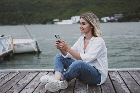 Charming young woman sits on pier on beautiful mountain lake and communicates on mobile phone