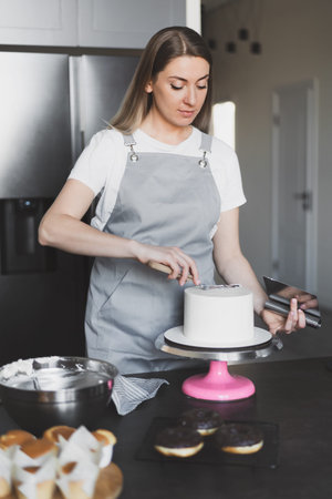 Young lovely woman pastry chef decorates cupcakes with whipped cream and having fun at home in...