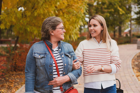 charming women in casual style, mom and daughter are sitting on bench in beautiful autumn park and looking at photos on phoneの写真素材