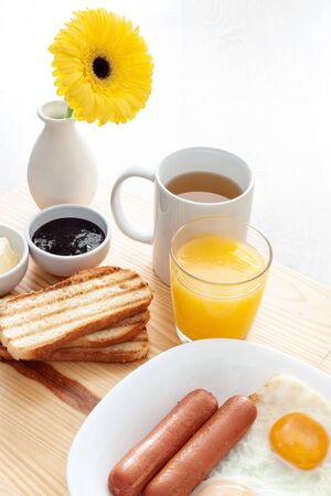 Breackfast with tea, juice, toasts, jam, butter, scrambled eggs, sausages and yellow gerber flower on wooden table macroの写真素材