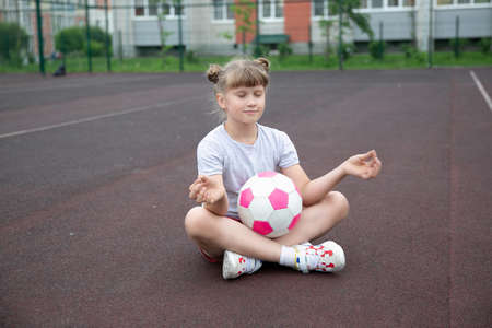 Young girl soccer player with a pink soccer ball isolated on a football field or brown background practicing yoga, meditatingのeditorial素材
