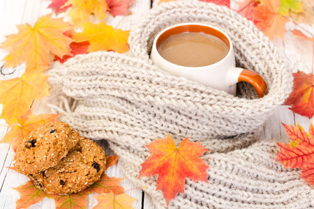 Cup of coffee and a warm scarf on a wooden background with autumn maple leavesの写真素材