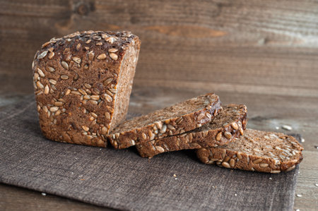 A loaf of fresh wholegrain bread cut into pieces on a brown wooden background.