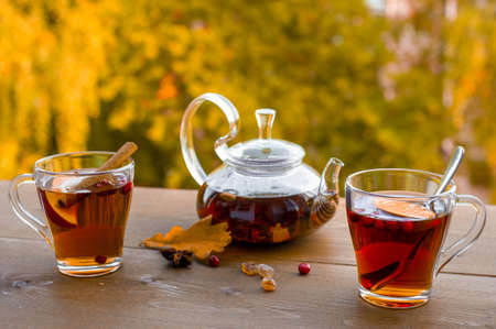 Autumn tea party outdoors. Glass kettle with hot herbal tea and two glass cups on a wooden table against the background of yellow trees.の写真素材