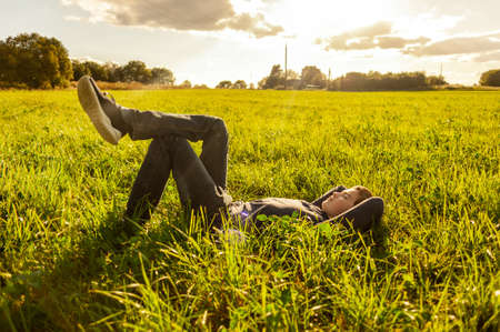 Rest in nature. A teenage boy lies on a green grass lawn in a rural area at sunset.の写真素材