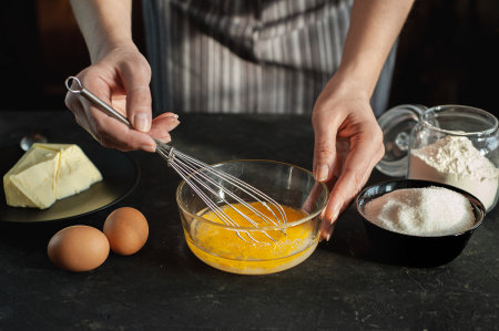 The process of preparing the dough. The woman whips the egg mixture with a whisk against a dark background. Ingredients for cookies.の写真素材