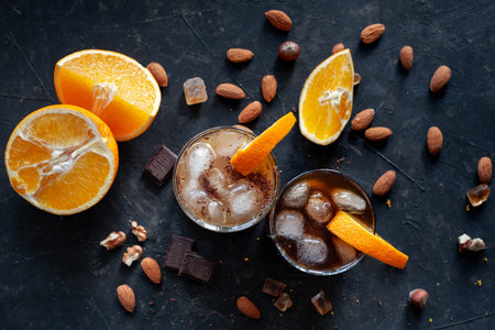 Cold espresso coffee with ice and orange juice in two glass glasses on a dark table with nuts,...