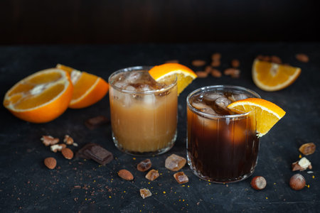Cold espresso coffee with ice and orange juice in two glass glasses on a dark table with nuts,...