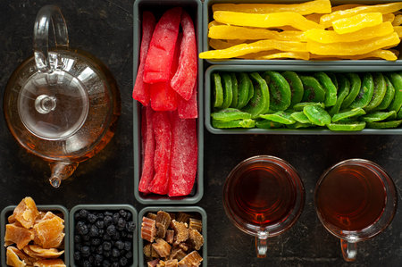 A set of different dried fruits and candied fruit in rectangular and square containers, two cups and a teapot with tea on a black background. Knolling. A healthy snack. Winter sweets. Vegan food.の写真素材