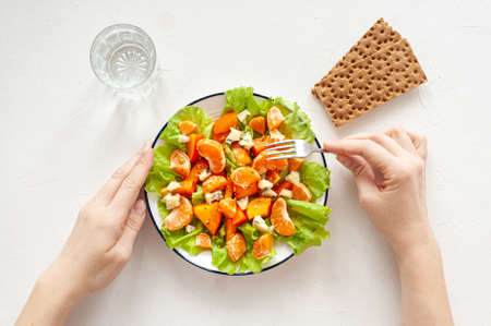 Bright vitamin salad with persimmon, tangerines and blue cheese on a white table. Womens hands hold a fork and a plate of salad. Dietary vitamin food. View from above.の写真素材