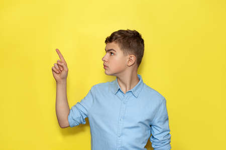 A teenage Caucasian boy in a blue shirt on a yellow background points his finger at something. A place for text.の写真素材