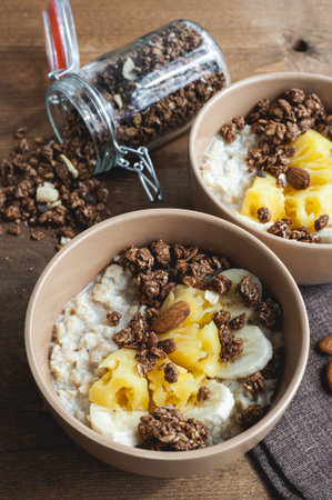 Oatmeal with pineapple, banana and chocolate granola in beige plates on a brown wooden table. A hearty healthy breakfast. A place for text.の写真素材