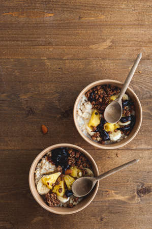 Oatmeal with pineapple, banana and chocolate granola in beige plates on a brown wooden table. A hearty healthy breakfast. A place for text.の写真素材