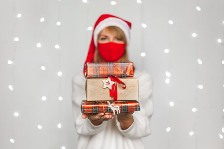 A beautiful blonde woman in Santas cap, red protective mask and white sweater holds boxes of Christmas gifts against the backdrop of festive lights. Christmas during the pandemic. Selective focusの写真素材