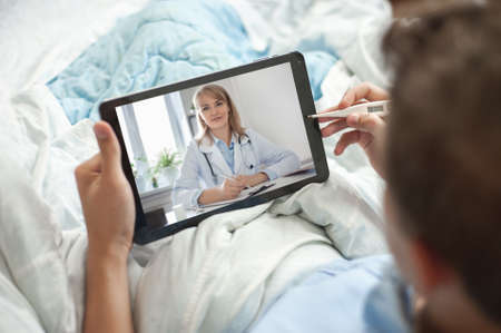 Telemedicine and online consultation concept. A young man or teenager sits in bed with an electronic thermometer and a tablet in his hands. The young man has video chat with a doctor.の写真素材