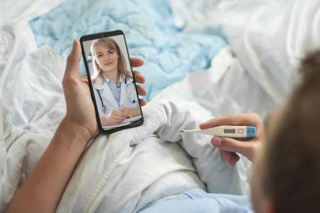 Concept of telemedicine and online consultations. A young man or teenager sits in bed with an electronic thermometer and a smartphone in his hands. A young man has a video chat with a doctor.の写真素材