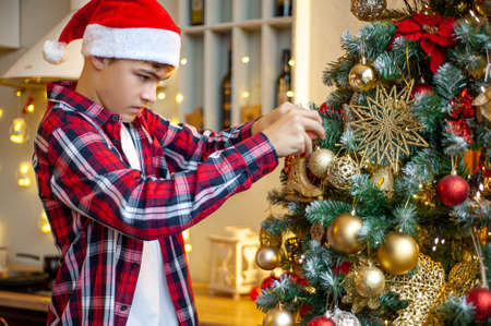 A Teenage Caucasian boy in a Santa hat decorates a Christmas tree in the home interior. Close-up and selective focus. Preparing for Christmas.の写真素材