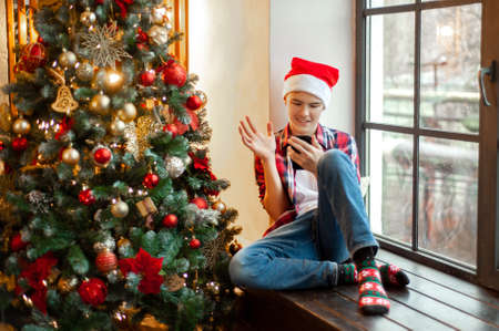 Teenage boy in Santas hat sits in a christmas-decorated room and chats online. A boy with a smartphone in his hand has a video chat and greets someone waving. Online communication at Christmas.の写真素材