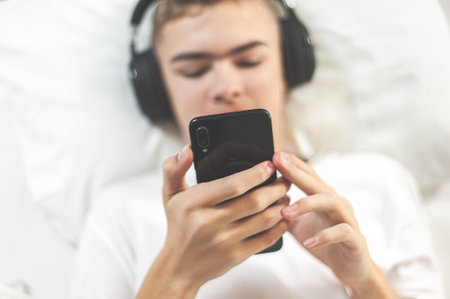 Teenager lies in bed with headphones and watches videos or listens to music on a smartphone Digital...