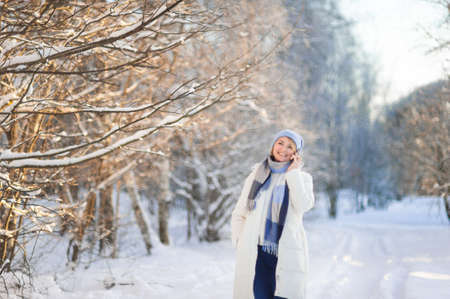 an attractive laughing blonde woman in a white coat and blue hat walks through a winter snow-covered park and talks on the phone. Active and healthy lifestyle.の写真素材