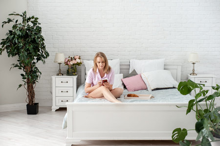 An attractive blonde woman in pajamas sits on a bed in the bedroom with a smartphone and a cup in...
