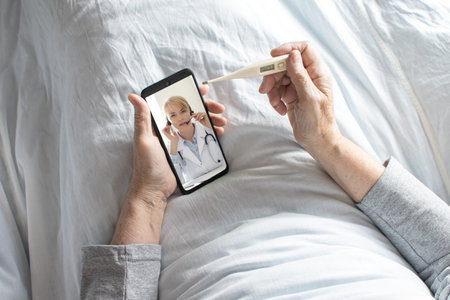 Elderly woman sitting in bed under a white blanket measures her temperature with an electronic...