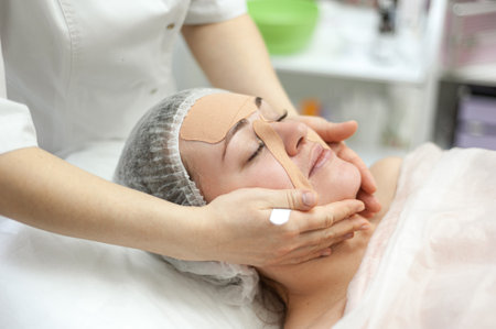 Professional care for the health and beauty of the face. Face taping. An attractive brunette woman lies on the couch at the dermatologist. The hands of the cosmetologist massage the patient.の写真素材