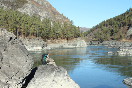 The traveler with a backpack sitting on a stone on the river bank among mountainsの写真素材