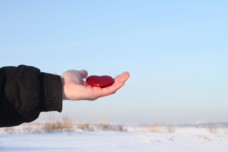 Symbolical red heart in a male palm against the blue sky and a snow fieldの写真素材
