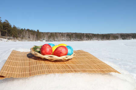 Multicolored Easter eggs in the wooden wattled plate standing on snow outdoorsの写真素材
