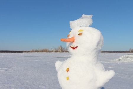 Snowman in a hat from ice standing on a snow glade in park against the blue skyの写真素材